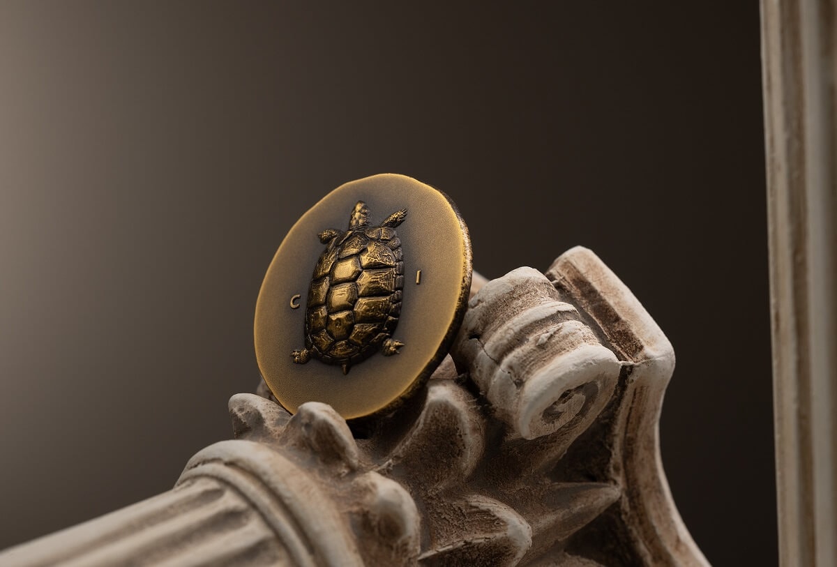 Golden turtle coin embedded on a white ornate column capital, symbolizing durability and longevity, with a simple 'C' and 'I' inscription.