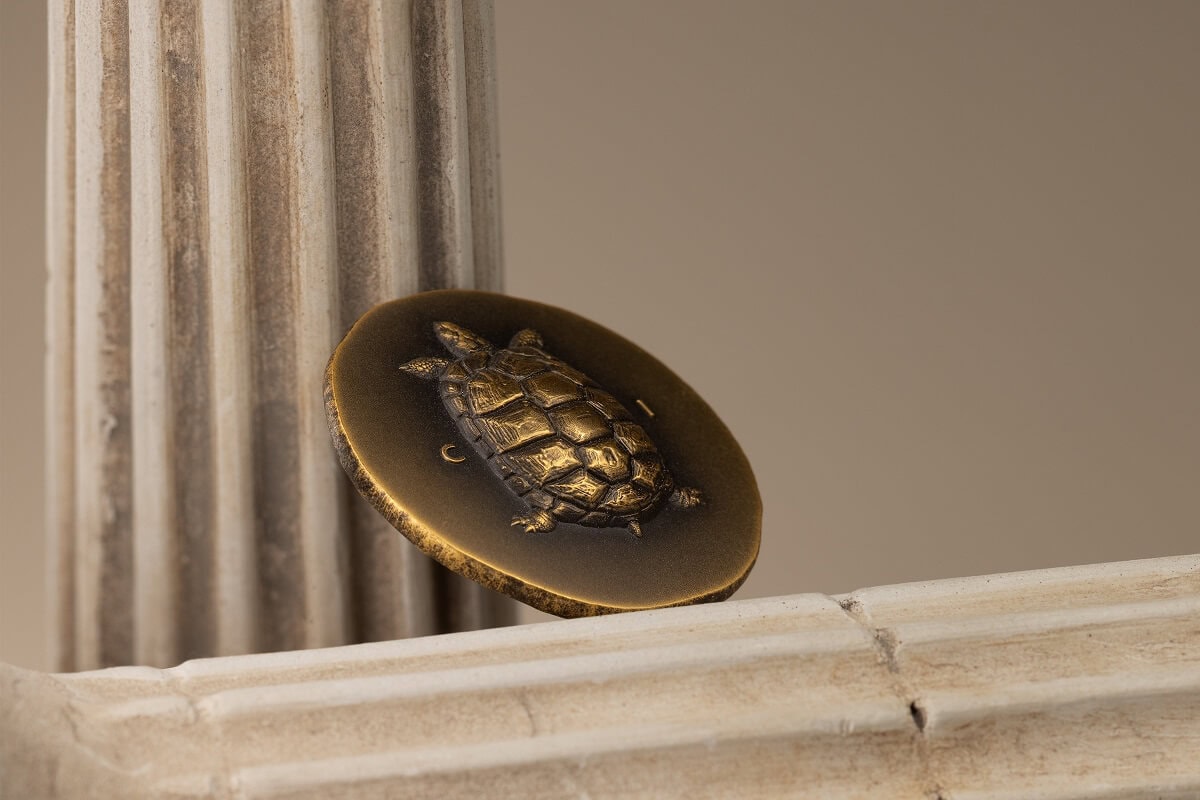 A collectible gold-toned turtle coin featuring detailed shell engravings, displayed on a marble shelf near a classical column.
