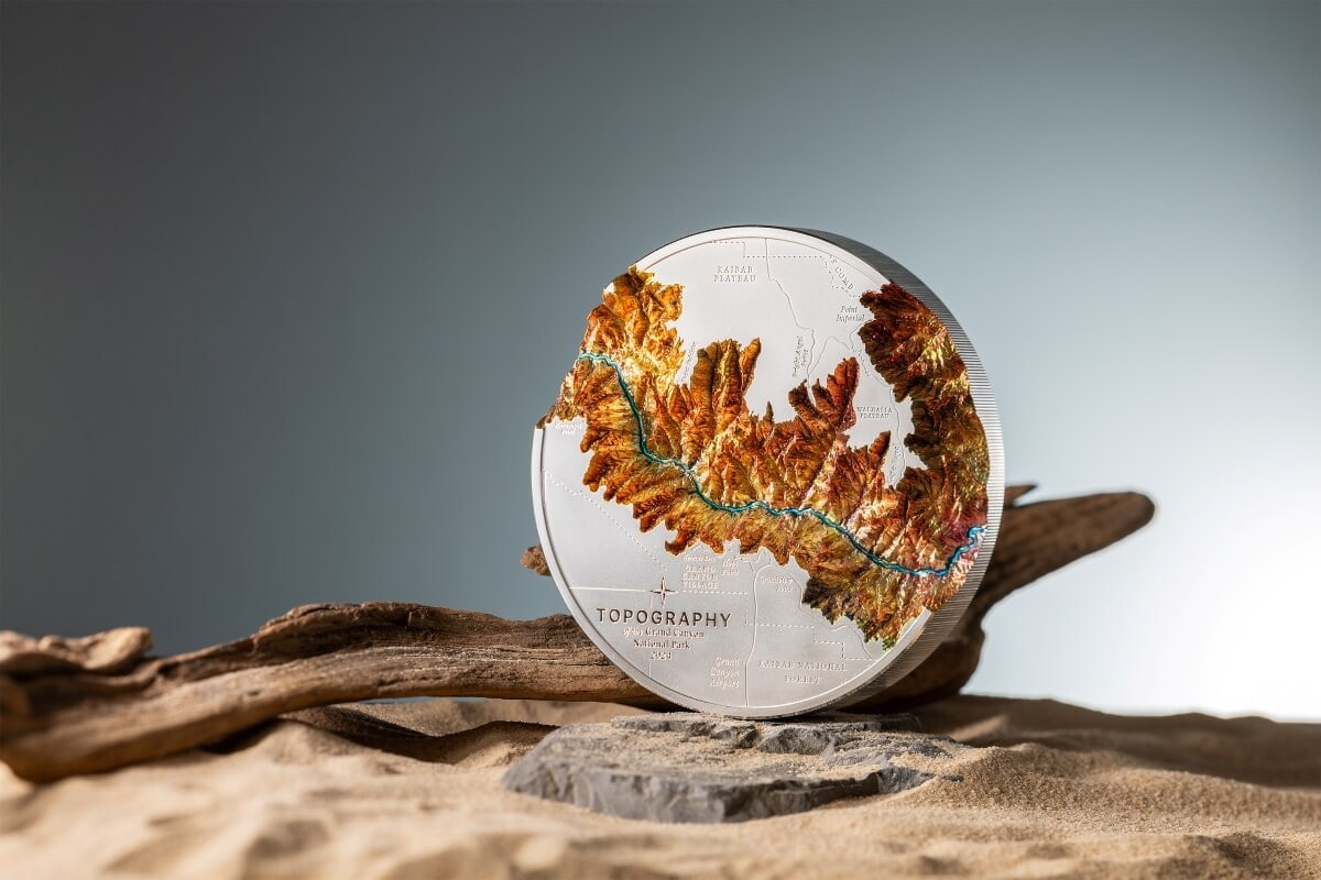 Detailed topography coin featuring colorful mountain terrain, river detail, labeled geographic features, and the text "TOPOGRAPHY" with additional inscriptions, set against a neutral background on driftwood.