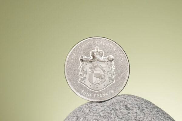 A 2024 Liechtenstein 100 Years of the Swiss Franc 1oz Silver Silk Finish Colorized Coin, standing upright on its edge on a light-colored, textured stone or rock, with a plain background.
