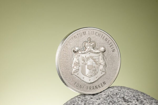 A 2024 Liechtenstein 100 Years of the Swiss Franc 1oz Silver Silk Finish Colorized Coin, displayed on what appears to be a light-colored, textured stone or rock, with a blurred natural background.
