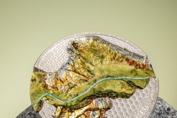 A 2024 Liechtenstein 100 Years of the Swiss Franc 1oz Silver Silk Finish Colorized Coin, placed on a light-colored, textured surface resembling stone or wood, with a blurred light green background. The coin's colorized map design is visible.