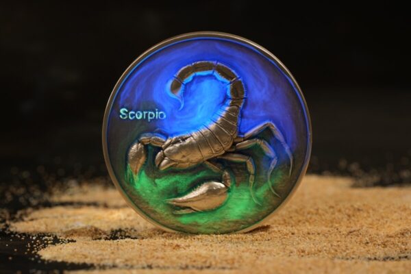 Aqua-colored Scorpio zodiac coin featuring a detailed scorpion engraving against a black background, resting on sandy surface.