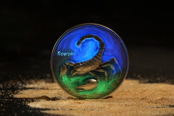 Scorpio zodiac coin with a detailed scorpion design, iridescent blue and green hues, placed on sandy surface.