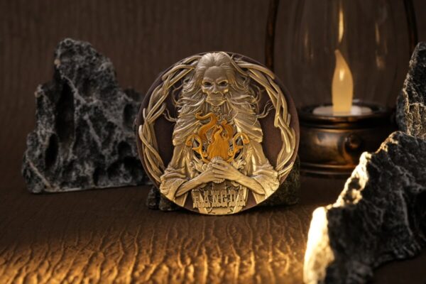 Gold-colored collectible coin featuring a lion-headed figure holding a flaming torch, with intricate designs and the word "RANFCA" at the bottom, displayed among black volcanic rocks and a candle holder.