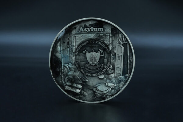 Asylum Cameroon 2025 silver collectible coin featuring a detailed design of an asylum with symbolic elements and inscriptions including the country name, denomination, and year.