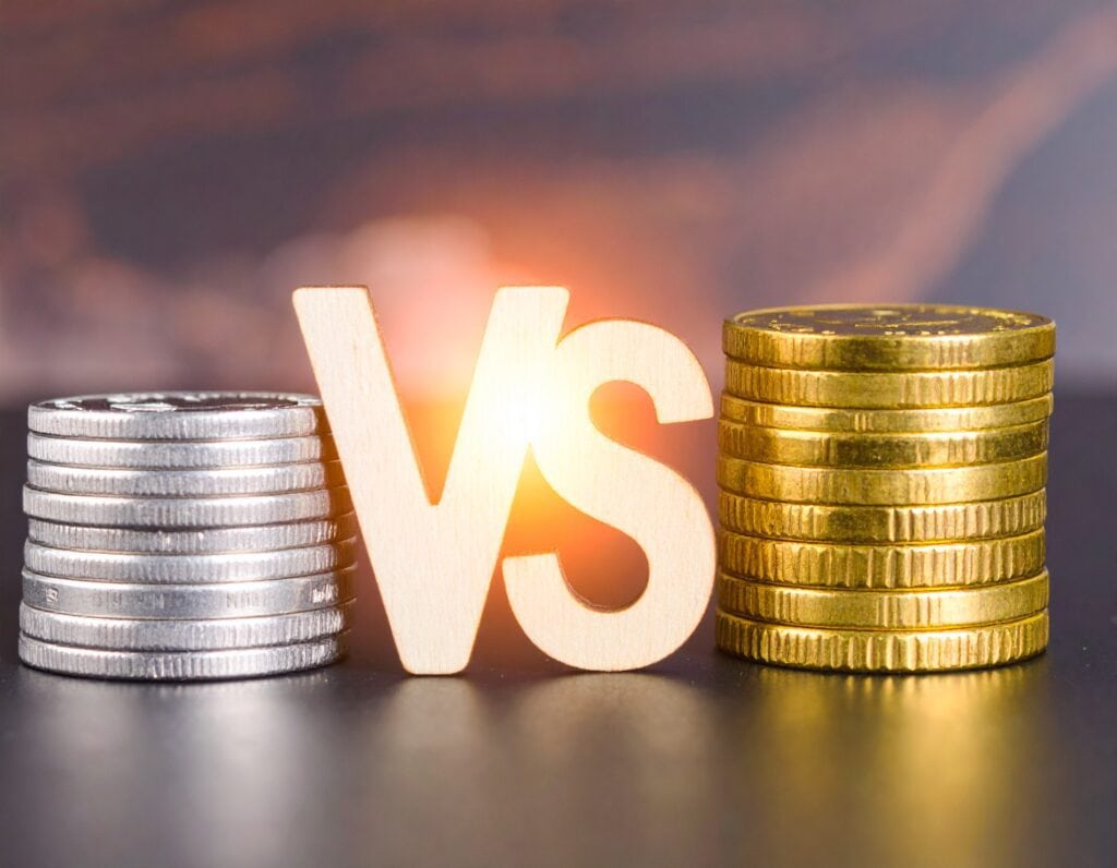 A stack of silver coins on the left, a large "VS" (versus) symbol in the center, and a stack of gold coins on the right, illuminated by warm light on a blurred financial background.