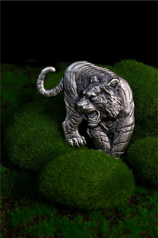 Lifestyle image of the 2025 South Korea Tiger 1 oz Silver Antiqued High Relief Shaped Medal placed on textured moss-green surface; medal features prowling tiger with open mouth, raised claw, and sculpted fur and muscle details in antiqued silver finish.