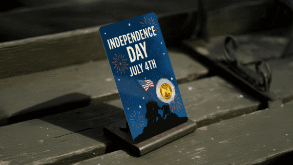 Front view of the 2026 Liberia United We Stand 1.1000 oz Gold Coin display card; shows the headline “INDEPENDENCE DAY JULY 26” with a dramatic illustration of soldiers raising a flag and fireworks above, with a circular gold coin embedded beneath the artwork, card placed on a stand.