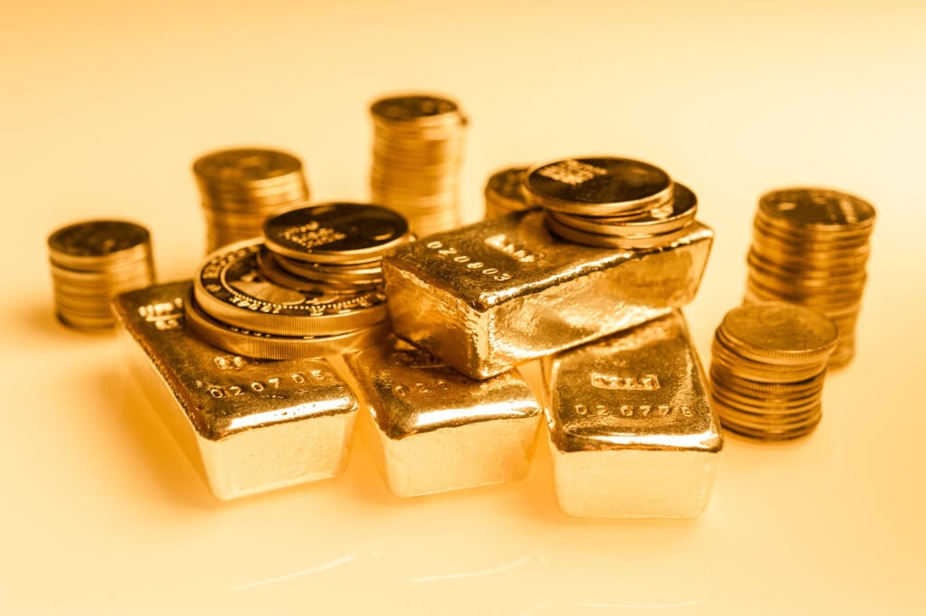 A collection of gold bullion bars and stacked gold coins, shown in a warm, golden light on a reflective surface.