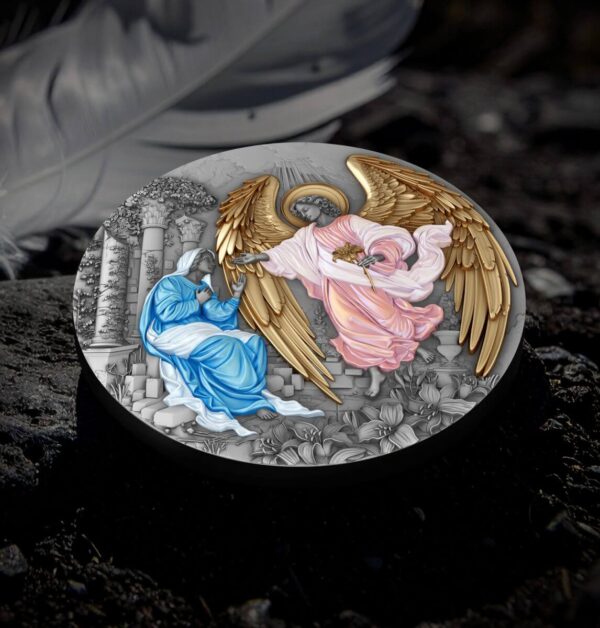 The front of a 2025 Niue "Herald of Salvation - Gabriel" antiqued and gilded silver coin, depicting the Annunciation, lying on a dark, textured surface with a blurred feather or leaf in the background.