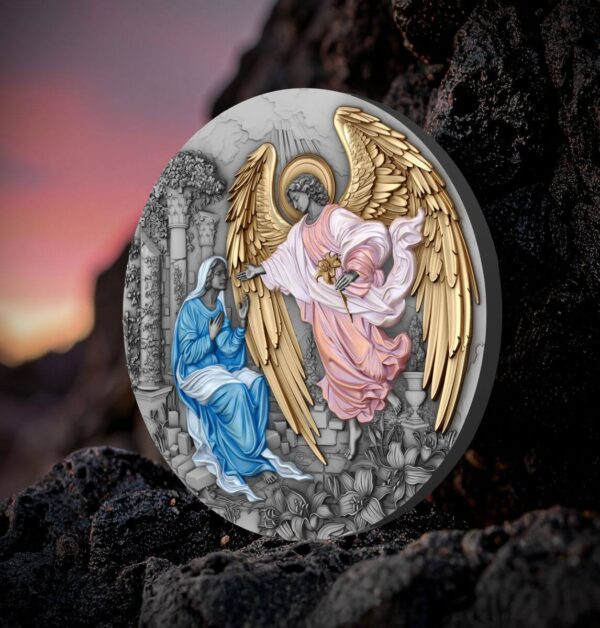 An angled view of the front of the Niue "Herald of Salvation - Gabriel" coin, showing the gilded Angel Gabriel and the colorized Virgin Mary, propped up on a dark, textured rock with a blurred sunset background.