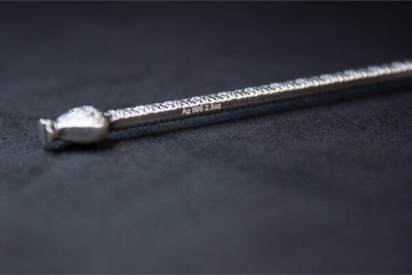 A close-up, side-view photograph of the bottom half of a silver, antiqued medal shaped like a winged staff, showing its thickness and a small mark.