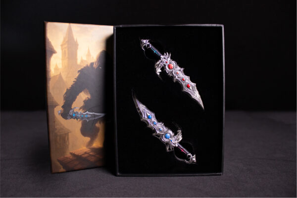 The Twinfang Blades coin set is shown inside a display box, with the two sword-shaped coins resting on a black velvet interior and the decorative sleeve next to the open box.