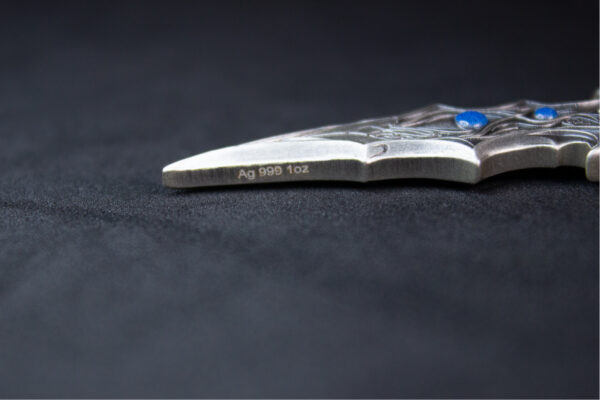 A close-up side view of an antiqued silver sword-shaped coin, showing its thickness and the texture of the blade and hilt with blue gems