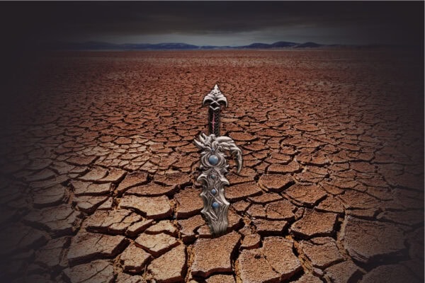 An antiqued silver sword-shaped coin with blue gems, standing upright in a cracked, dry desert landscape under a cloudy sky.