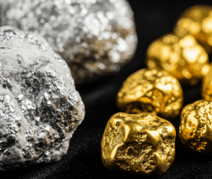A close-up, studio shot of two piles of raw metal nuggets on a black background: one pile of raw, silvery platinum nuggets and one pile of shiny, raw gold nuggets.