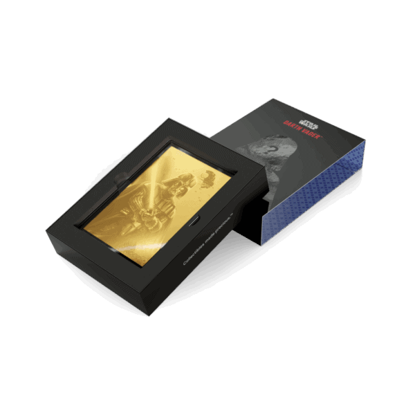 A gold-colored, rectangular Star Wars Darth Vader coin is displayed in a black box with a clear lid. A separate, dark blue and black box with a graphic of Darth Vader is next to it