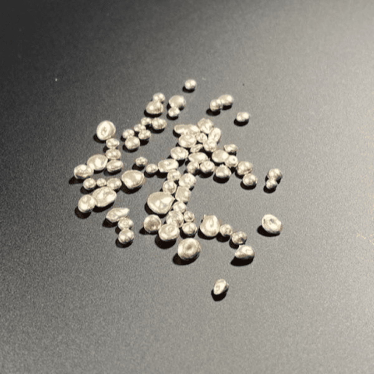 Unfocused close-up of lustrous white pearls scattered on a dark matte surface, capturing their irregular shapes and reflective surfaces.